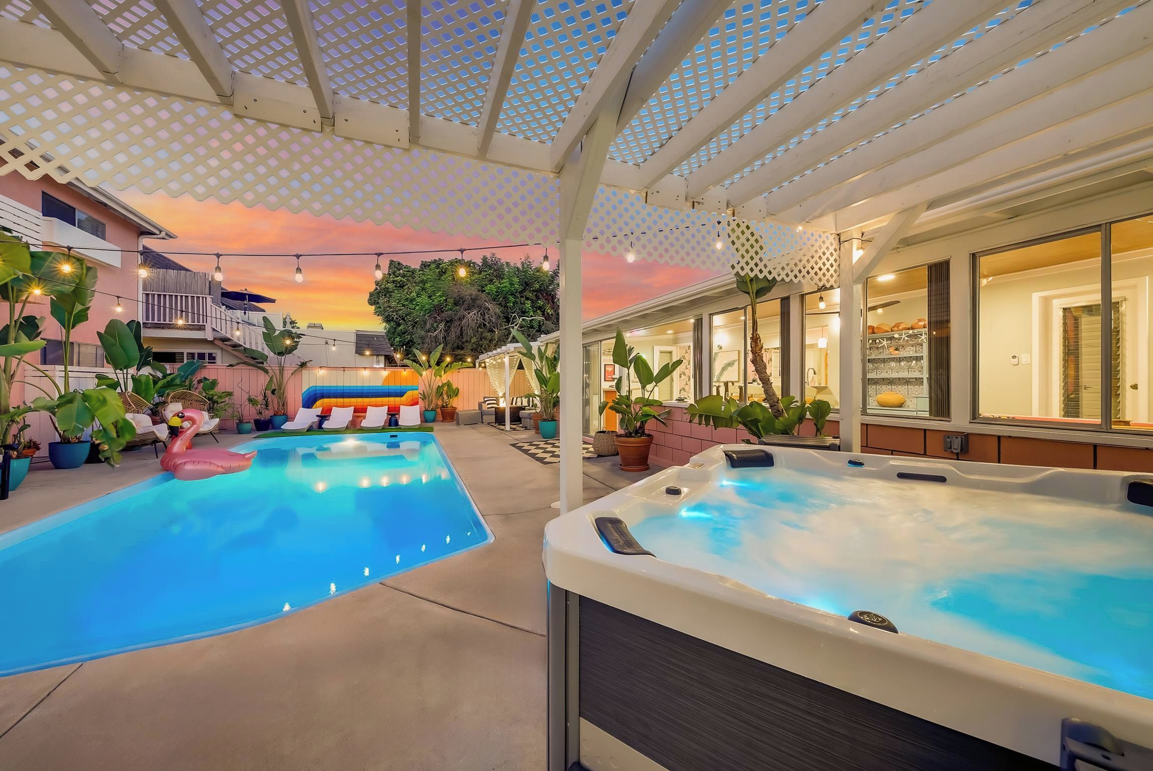 A pool area with a hot tub, lounge chairs, and a covered patio with a view of a building.