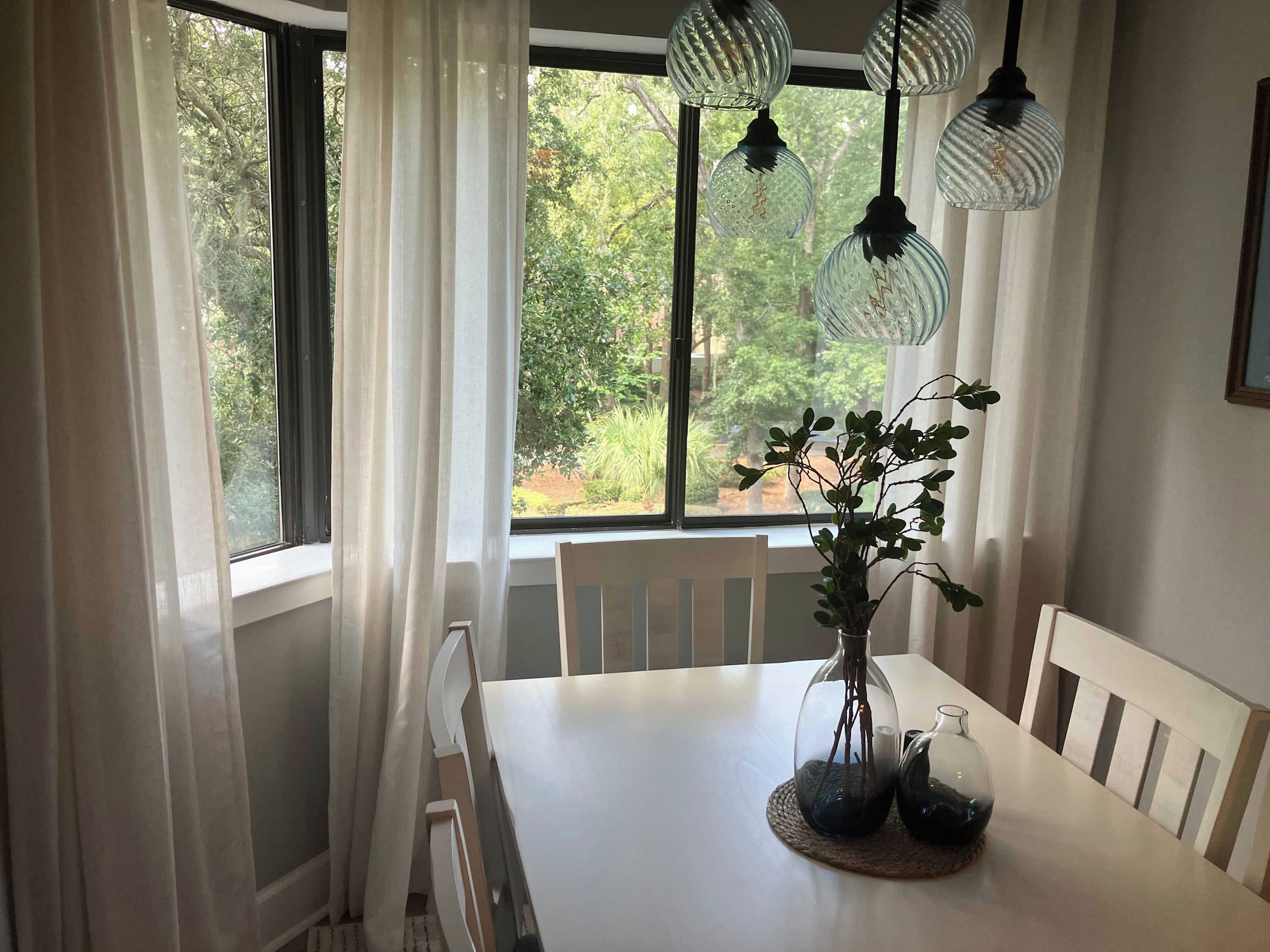 A dining area with a table, chairs, a vase with plants, and hanging light fixtures.