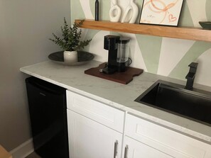 Una cocina con encimera blanca, cafetera, planta en maceta y estante con adornos.