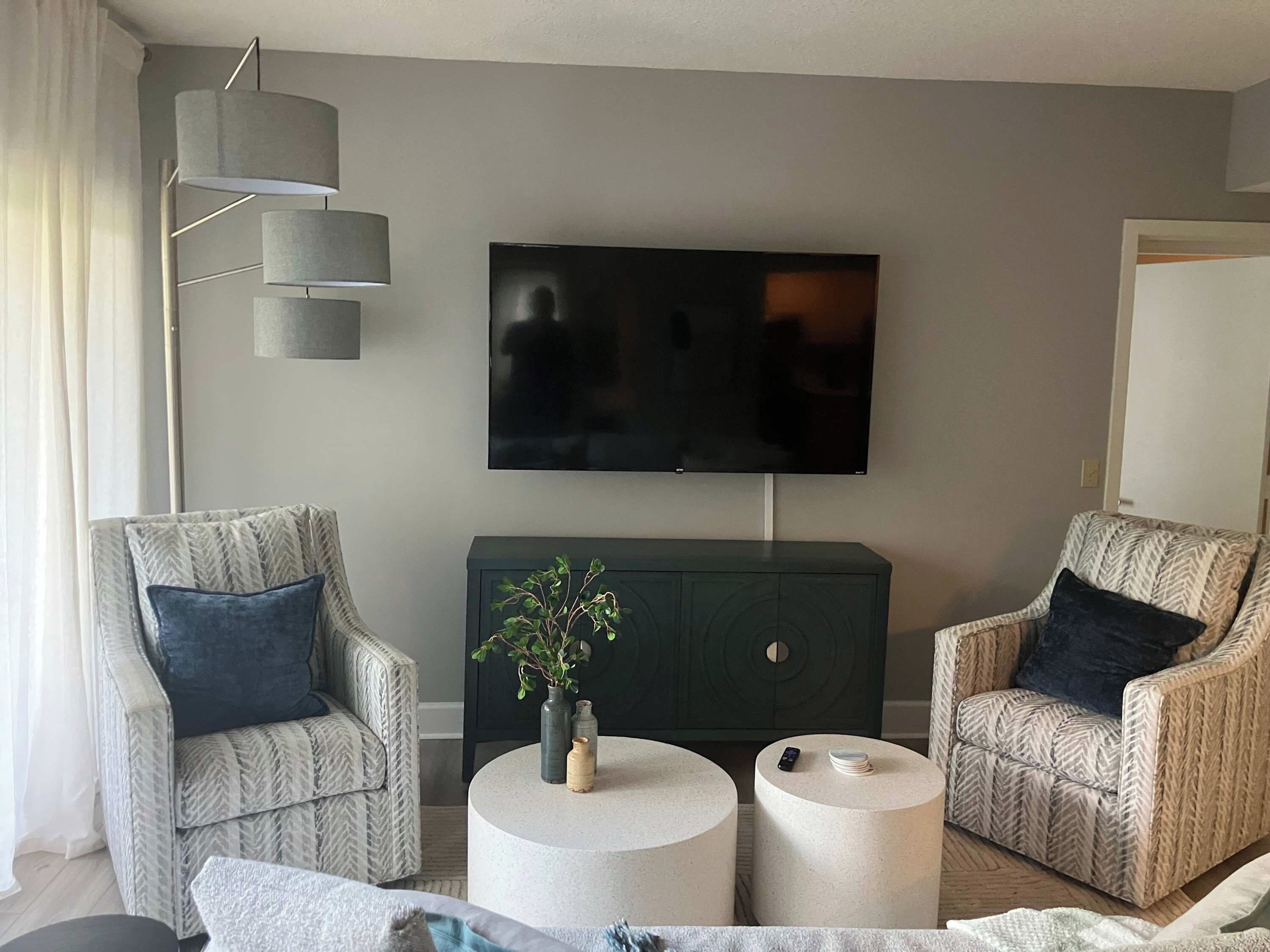 Ein modernes Wohnzimmer mit zwei gemusterten Sesseln, einem runden Couchtisch, einem an der Wand befestigten Fernseher und einem dunklen Schrank.