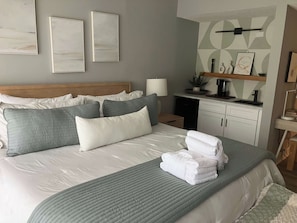 A hotel room with a bed, pillows, a bedside table, a lamp, a small plant, a coffee machine, and a wall with abstract art. - Village House 204 | Relax & Walk to the Beach (Hilton Head Island)
