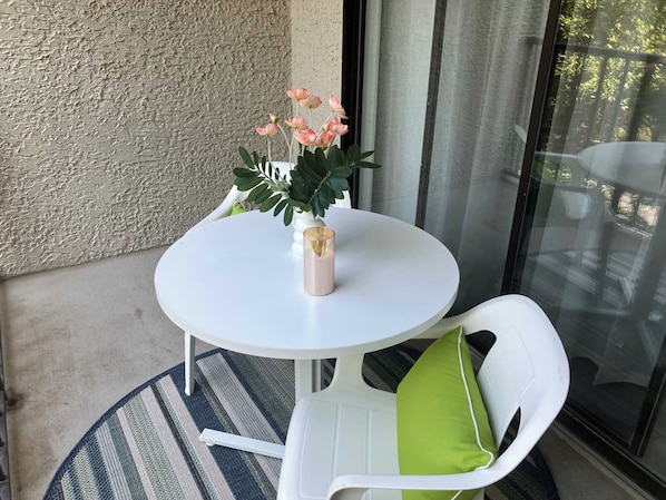 A small round table with a vase of flowers, a chair with a green cushion, and a textured wall in the background. - Village House 204 | Relax & Walk to the Beach (Hilton Head Island)