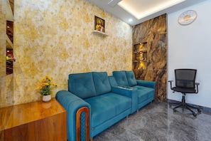 A reception area with a blue sofa, a wooden reception desk, a black office chair, and a marble wall with a clock. - Deluxe Apsara Tourist Homes (Adoor)