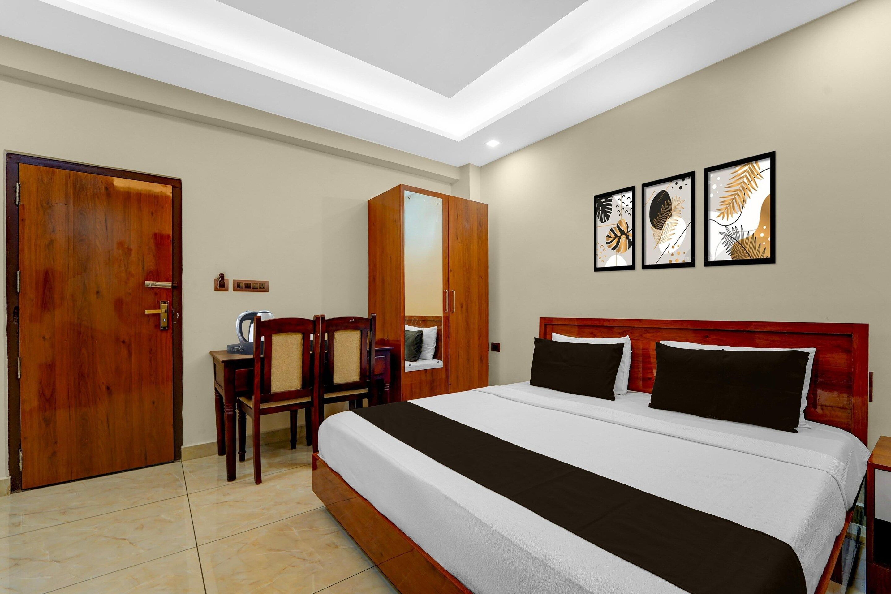 A hotel room with a large bed, wooden wardrobe, desk with chair, and three framed artworks on the wall.