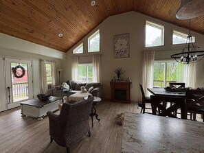A spacious living room with a high ceiling, wooden flooring, a fireplace, a dining area with a table and chairs, and large windows allowing natural light. - Whispering Woods 2bdrm, 2 bath, tucked in the woods. (Pittsfield)