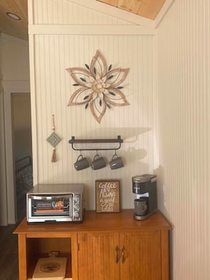 A kitchen area with a wall-mounted flower decoration, a coffee maker, and a toaster oven on a wooden cabinet. - Hummingbird is a Cozy & Quaint 1 bedroom Cabin bordering Ozark National Forest (Dover)