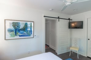 A bedroom with a bed, a painting, a ceiling fan, a television, and a chair.