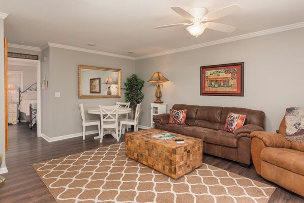 A living room with a brown leather sofa, a wooden coffee table, a white dining set, and a ceiling fan.