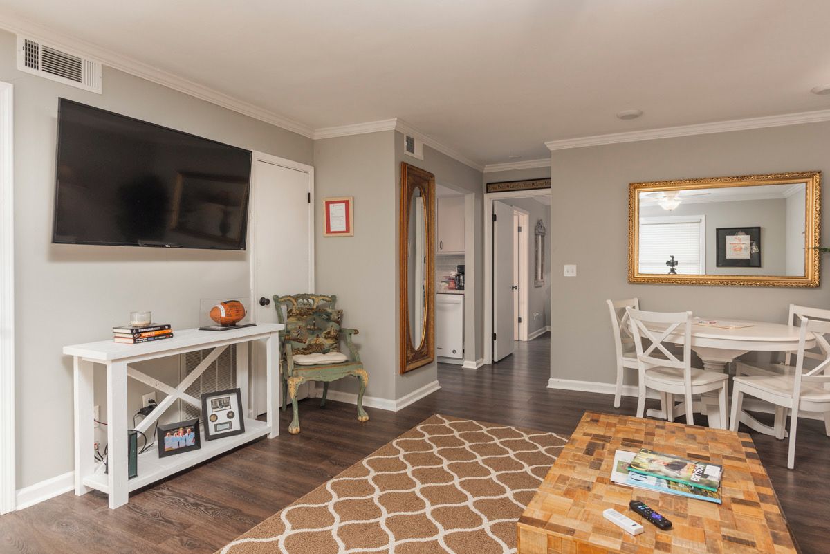 A living room with a television, a coffee table with remote controls, a patterned rug, and a dining area with chairs.