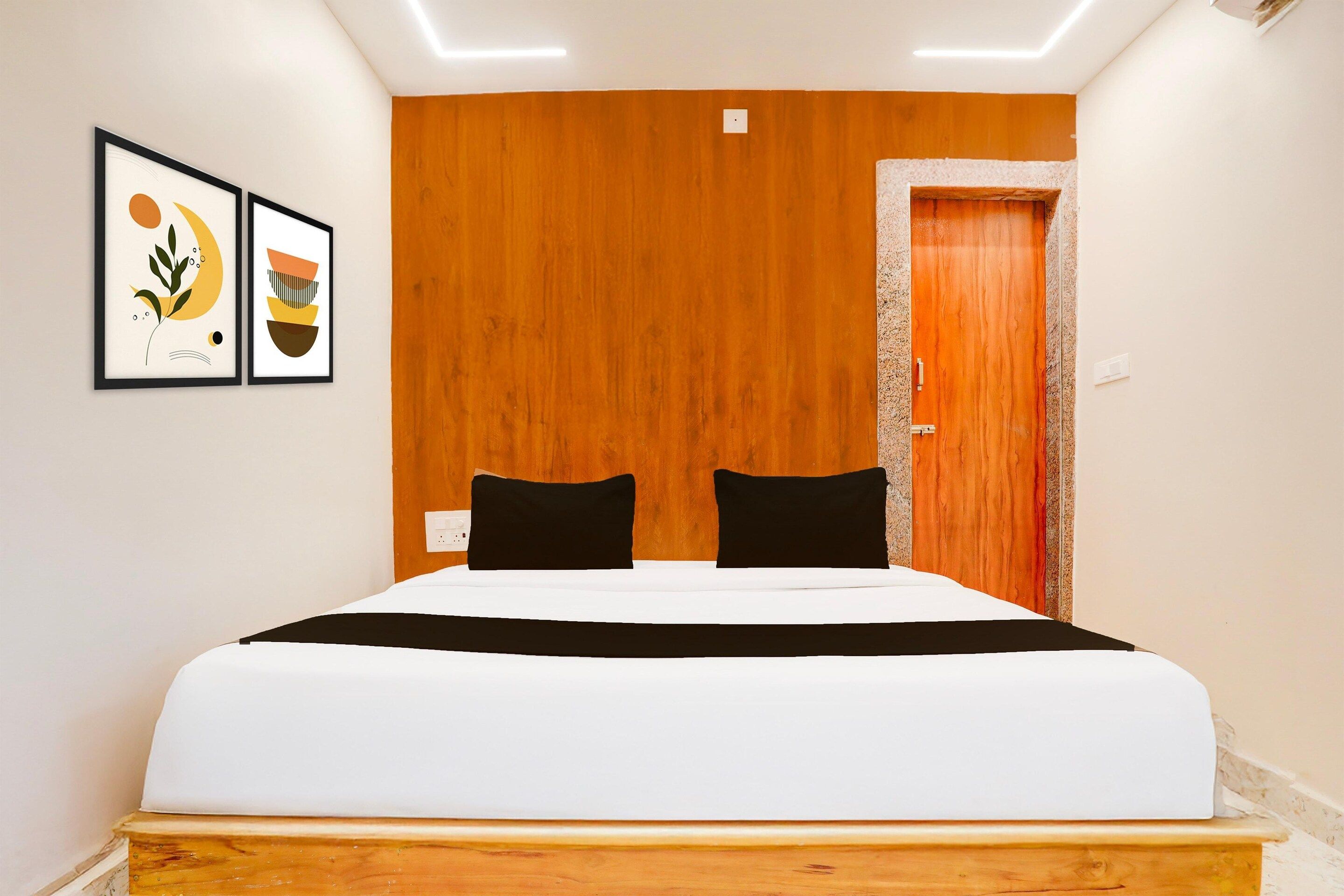 A hotel room with a wooden headboard, a bed with white bedding and black pillows, and two framed artworks on the wall.