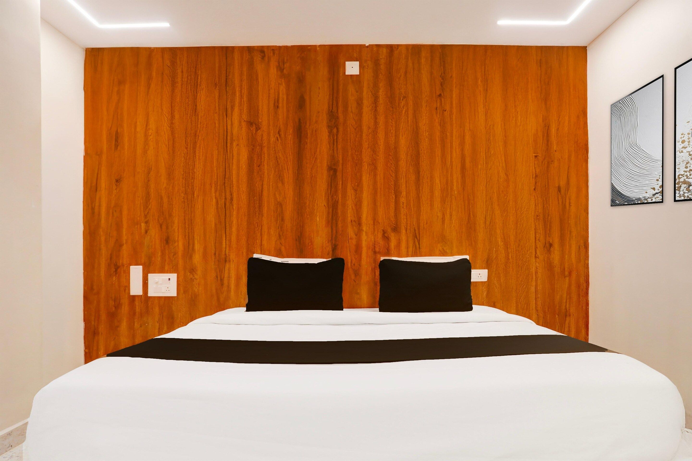 A hotel room with a wooden headboard, two black pillows, a white bedspread, and two wall-mounted light switches.