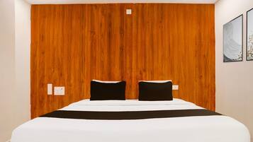 A hotel room with a wooden headboard, two black pillows, a white bedspread, and two wall-mounted light switches.