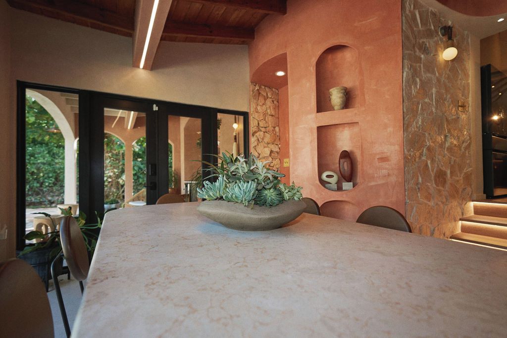 Comedor con mesa de mármol, sillas, un centro de mesa con planta y pared de piedra con estantes.