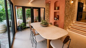 A modern dining area with a long table, chairs, and a bowl of plants. There are potted plants on the floor and a stone wall with shelves. - Vrbo Property (Miami)