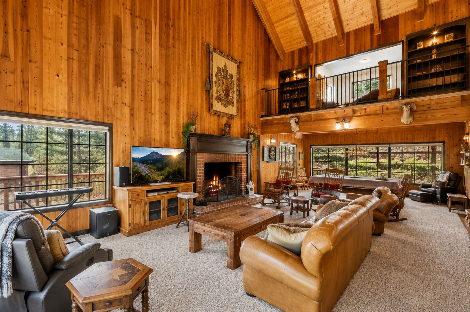 A spacious log cabin interior with a fireplace, leather sofas, a television, and a large window overlooking a forested area.