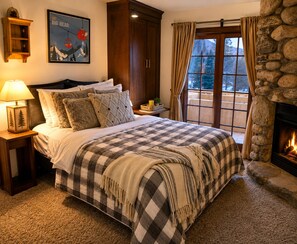 Quarto com cama grande, mesa de cabeceira com abajur, lareira de pedra e janela com cortinas.
