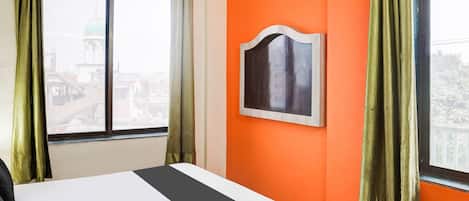 A hotel room with a bed, an orange wall, a window with curtains, and a framed picture.