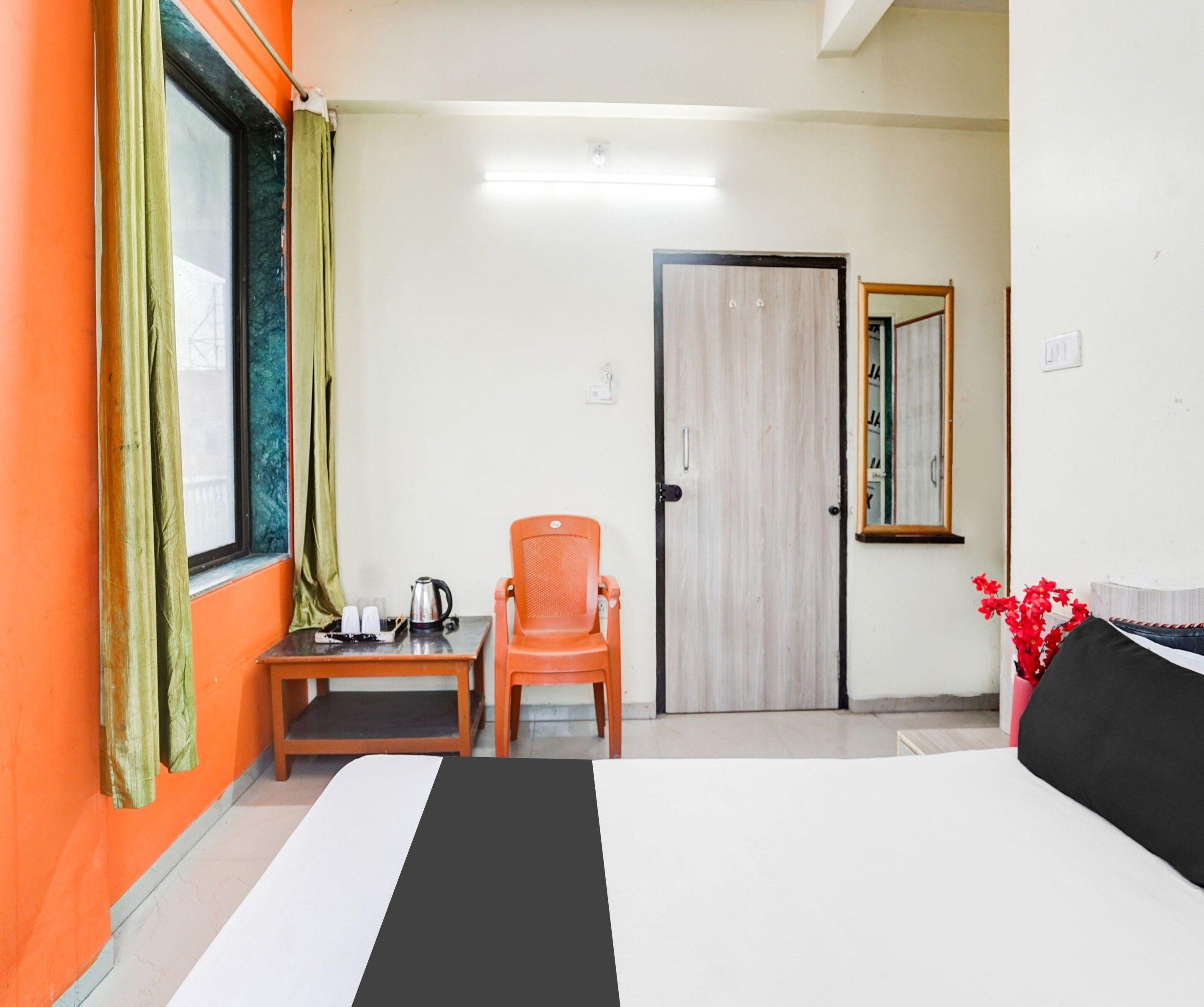 A hotel room with a bed, a chair, a small table with a kettle, a door, and a mirror.