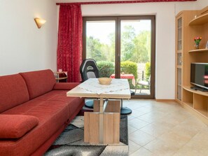 Ein Wohnzimmer mit einem roten Sofa, einem kleinen Tisch mit einem Korb, einem Fernseher auf einem Holzständer und einer Glastür zum Garten.