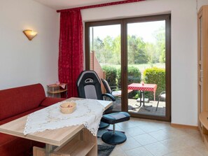 Ein Zimmer mit einem roten Sofa, einem Tisch mit weißer Tischdecke, einem Stuhl, einem kleinen Tisch mit roter Tischdecke und einer Glastür zu einem Außenbereich mit Tisch und Stühlen.