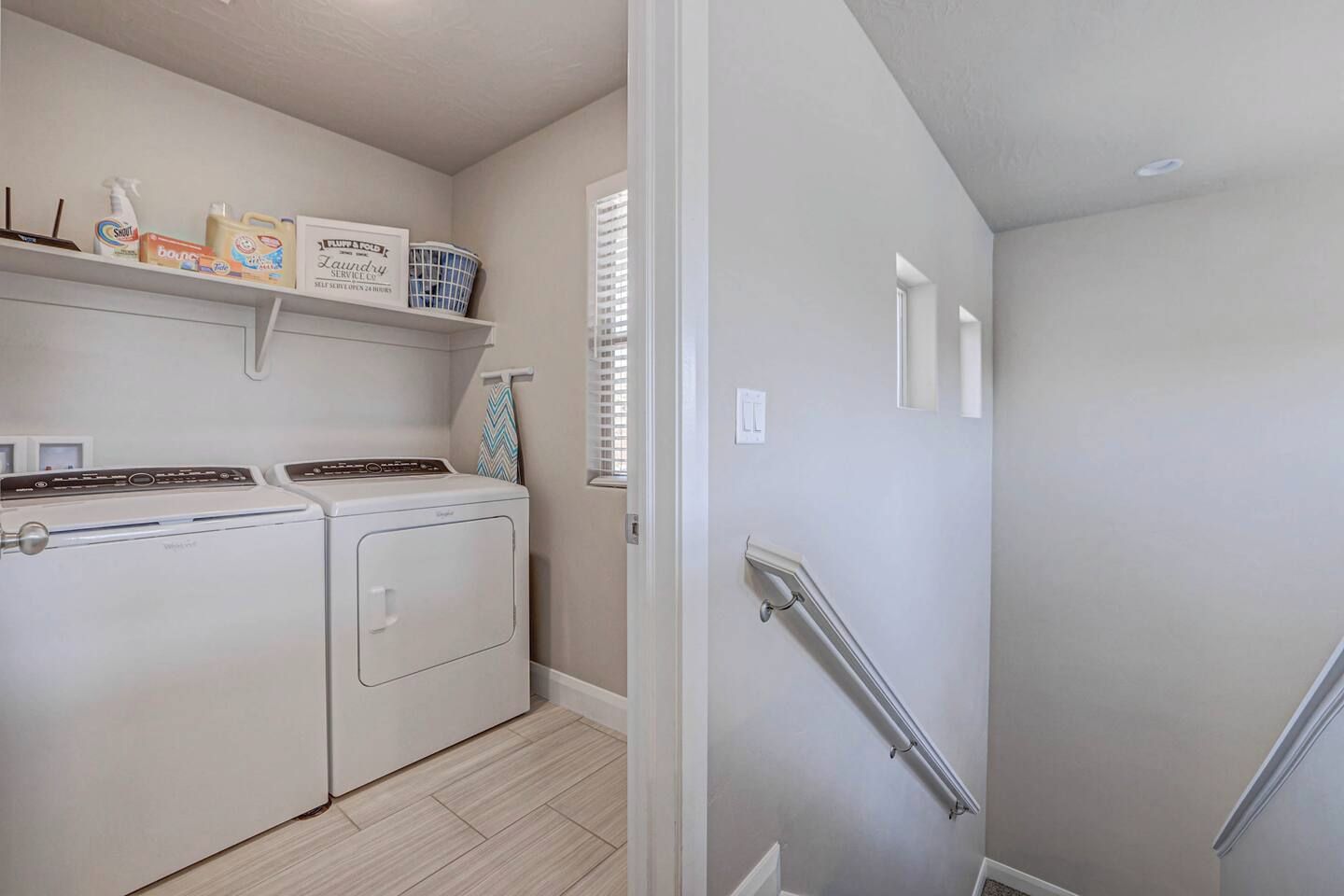 A laundry room with a washing machine and dryer, shelves with cleaning supplies, and a handrail on a staircase.