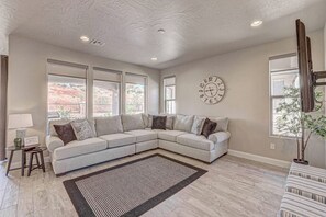 A living room with a sectional sofa, a clock on the wall, and large windows. - Hot Tub, Heated Pool, Ping Pong, Foosball, xBox, FastWiFi in Southern Utah (Washington)