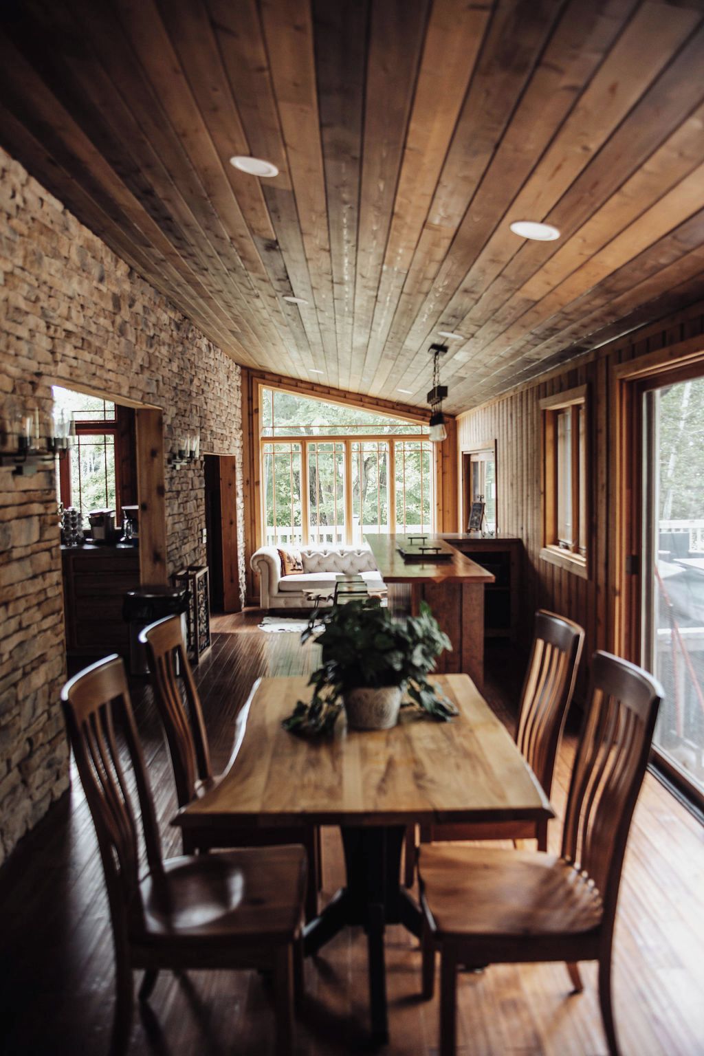 A wooden dining table with chairs, a potted plant, and a kitchen area with a stone wall and large windows.
