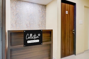 A reception desk with a sign indicating it's a company-serviced collection point, a wooden door with a number, and a wall with a floral pattern. - Deluxe Zudio 7 Baner (Pune)