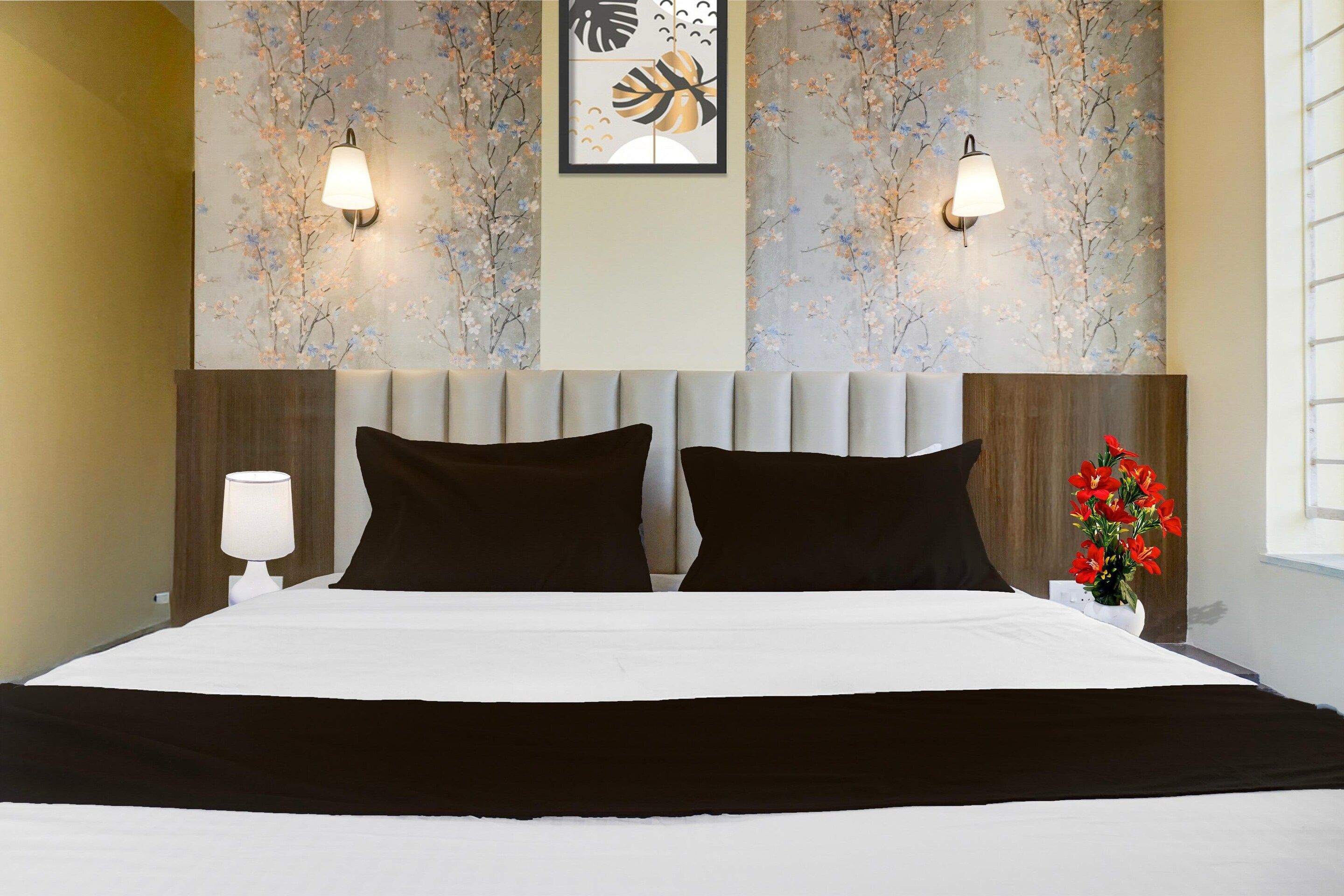 A hotel room with a bed, two wall-mounted lamps, a framed artwork, and a vase with flowers.