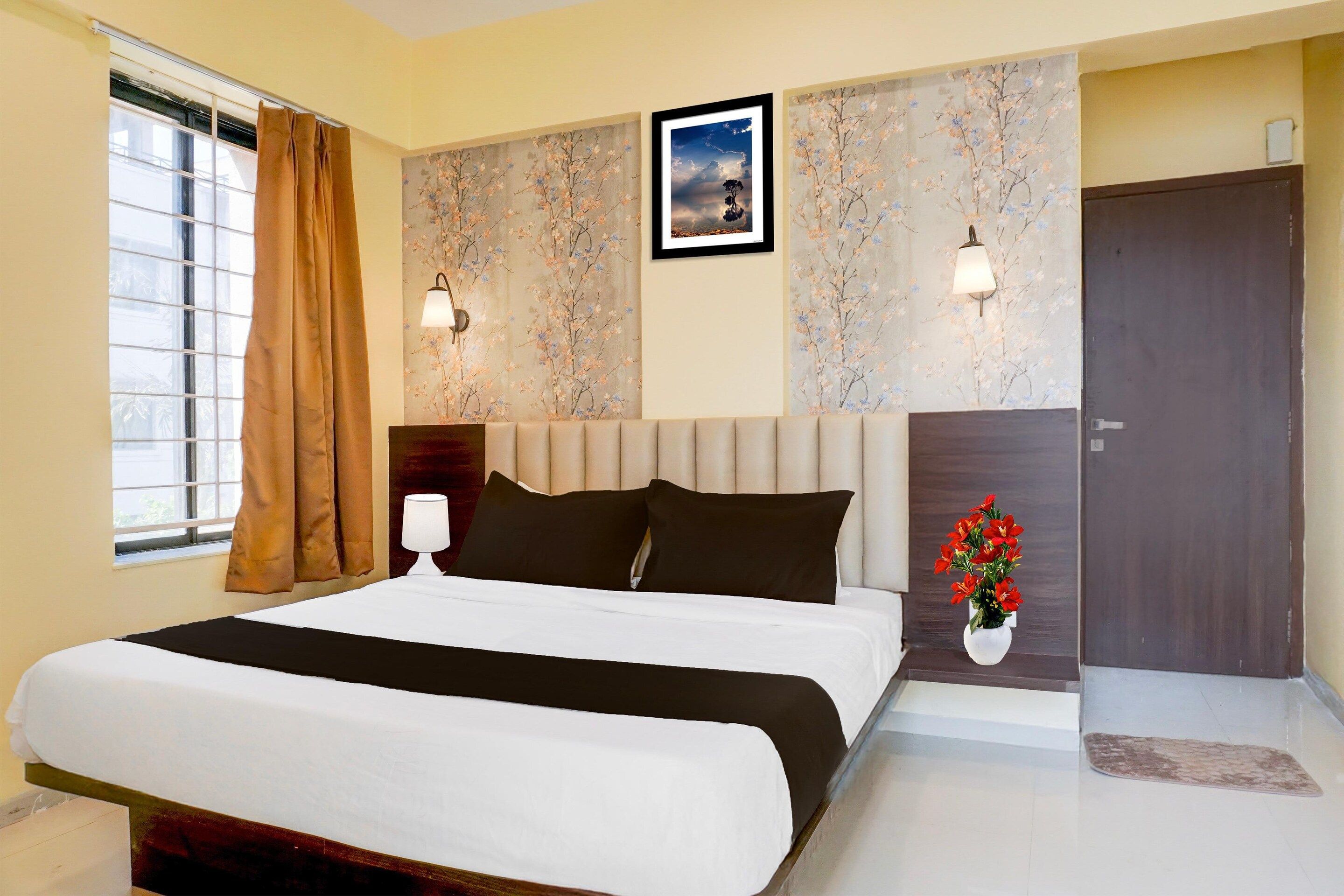 A hotel room with a bed, a nightstand, a lamp, a window with curtains, a door, and a wall with a framed picture.