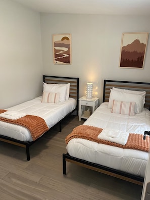 Habitación de hotel con dos camas individuales, una mesita de noche con lámpara y dos cuadros en la pared.