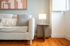 En stue med en grå sofa, mønstrede puder, et lille rundt bord med en lampe og et indrammet kunstværk på væggen.