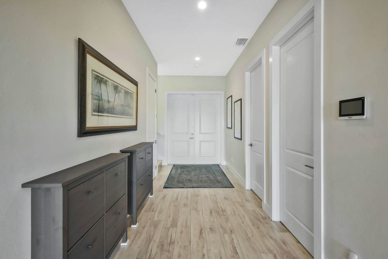 A hallway with a dark wooden dresser, a framed picture, a white door, and a small electronic device on the wall.