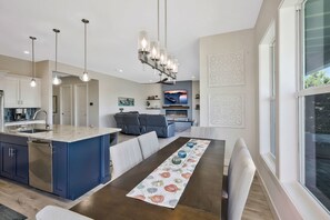 Eine moderne Küche mit einer zentralen Kochinsel, ein Essbereich mit Tisch und Stühlen sowie ein Wohnzimmer mit Sofa und Kamin.