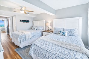 Quarto com duas camas, um ventilador de teto, mesa de cabeceira e vista para o mar.