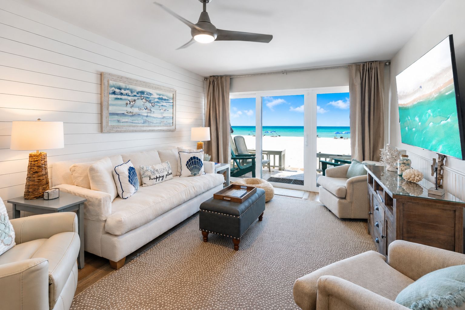 A spacious living room with a large window overlooking a beach, a ceiling fan, and a flat-screen TV.