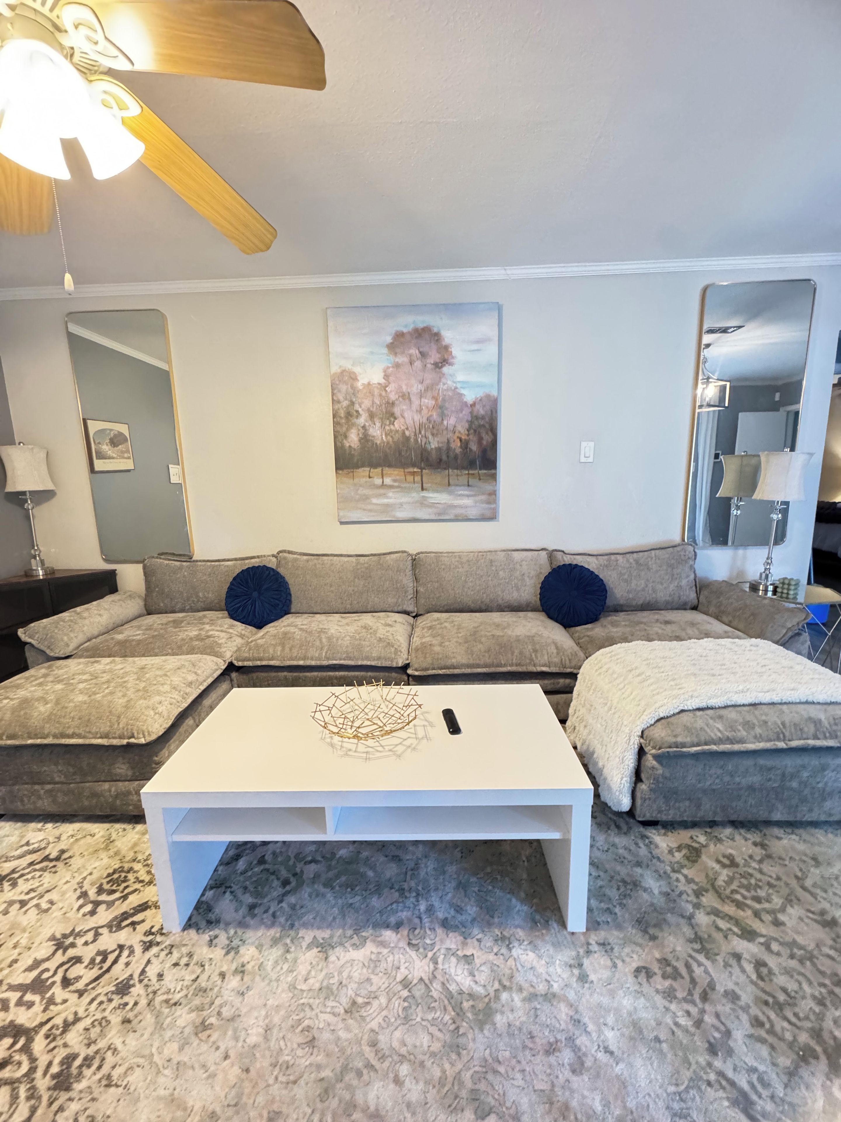 A living room with a sectional sofa, a white coffee table, a painting, and a ceiling fan.