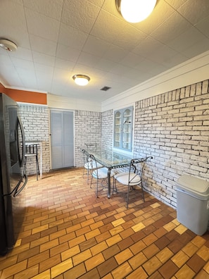 Una habitación con suelo de ladrillo, una mesa con cubierta de vidrio y sillas de metal, un refrigerador y un cesto de basura.