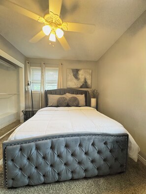 Un dormitorio con una cama grande, un ventilador de techo, una ventana con cortinas y una obra de arte colgada en la pared.