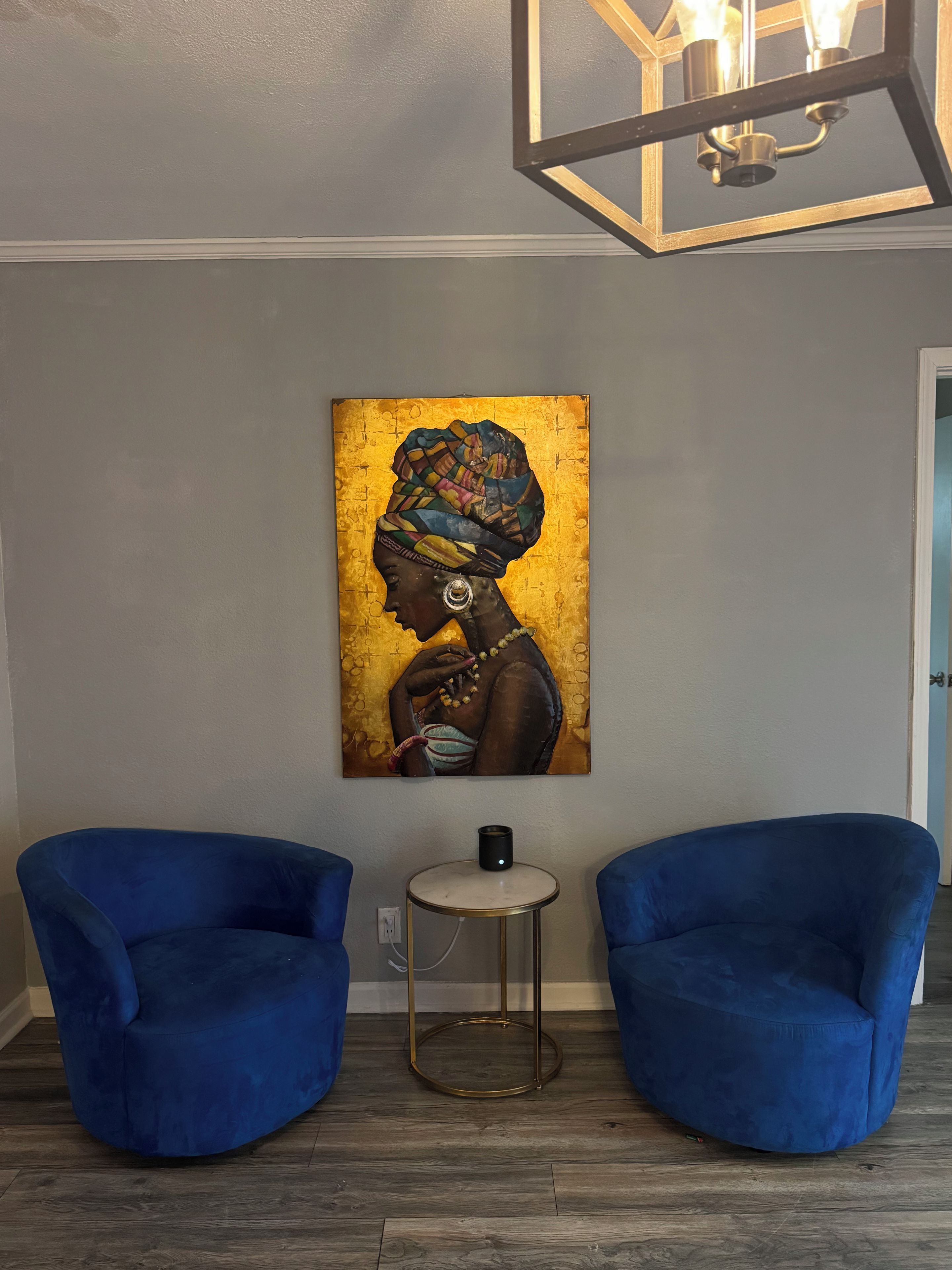 Une pièce avec deux fauteuils bleus, une petite table ronde avec une lampe et un tableau accroché au mur.