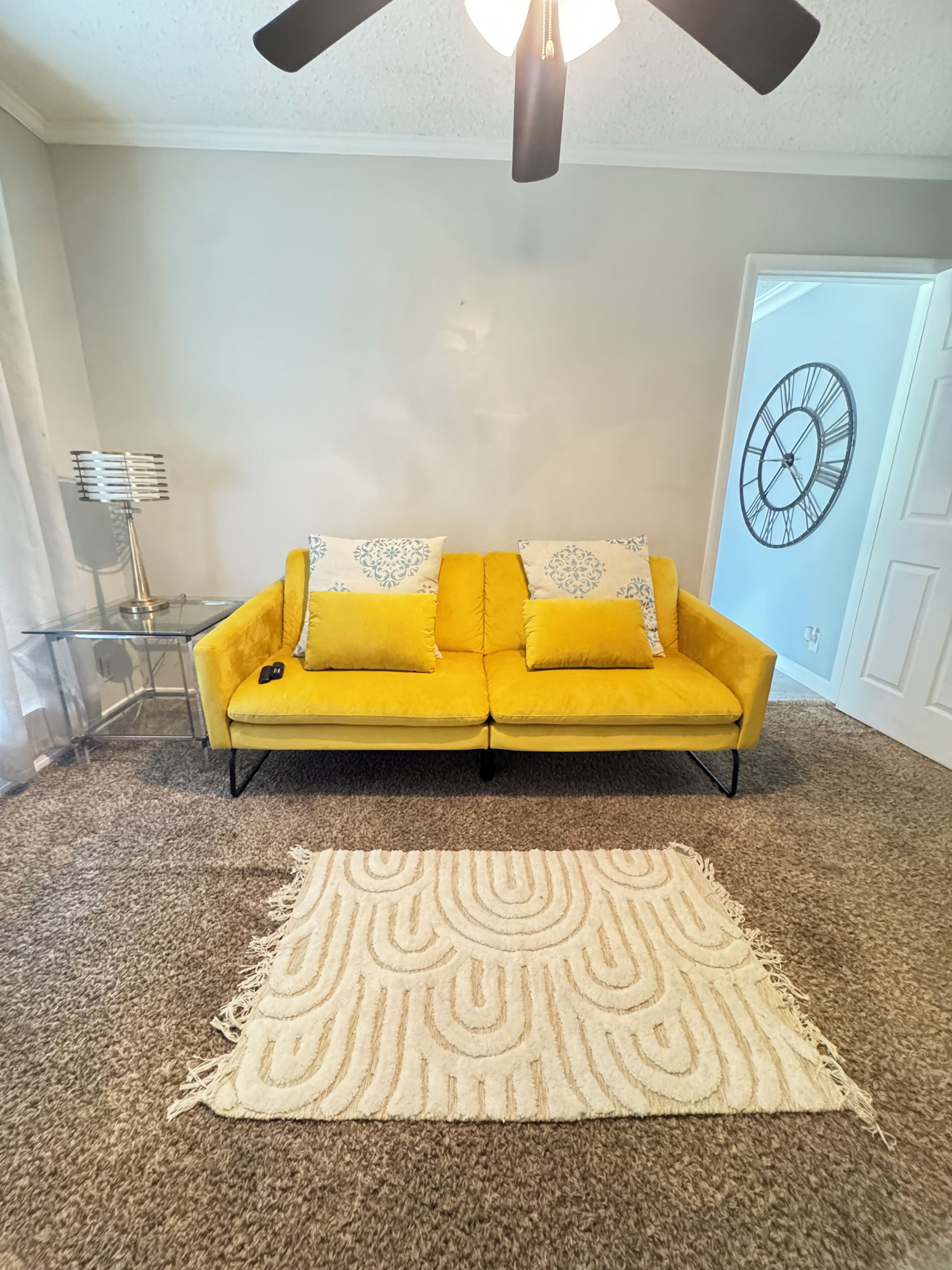 Une pièce avec un canapé jaune, un tapis à motifs, une horloge au mur et un ventilateur de plafond.