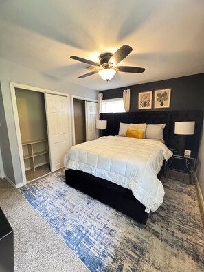 Un dormitorio con cama, ventilador de techo, ventana con cortinas, mesita de noche y lámpara de pared.