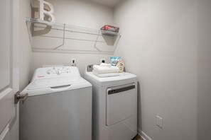 Un espace buanderie comprenant un lave-linge et un sèche-linge à chargement frontal, une étagère au-dessus du sèche-linge et un sac de lessive.
