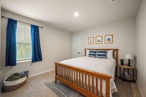 Un dormitorio con una cama de madera, una mesita de noche con lámpara, una alfombra pequeña y un ventanal con cortinas.