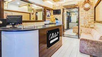 Uma mesa de recepção com uma placa indicando 'Reception', uma placa com o aviso 'Horário de Check-out: 12h' e uma placa informando 'Não é permitido trazer comida de fora'.