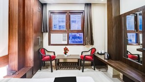 Habitación de hotel con un ventanal grande, un armario de madera, una mesita con sillas y una mesita de noche con una tetera.