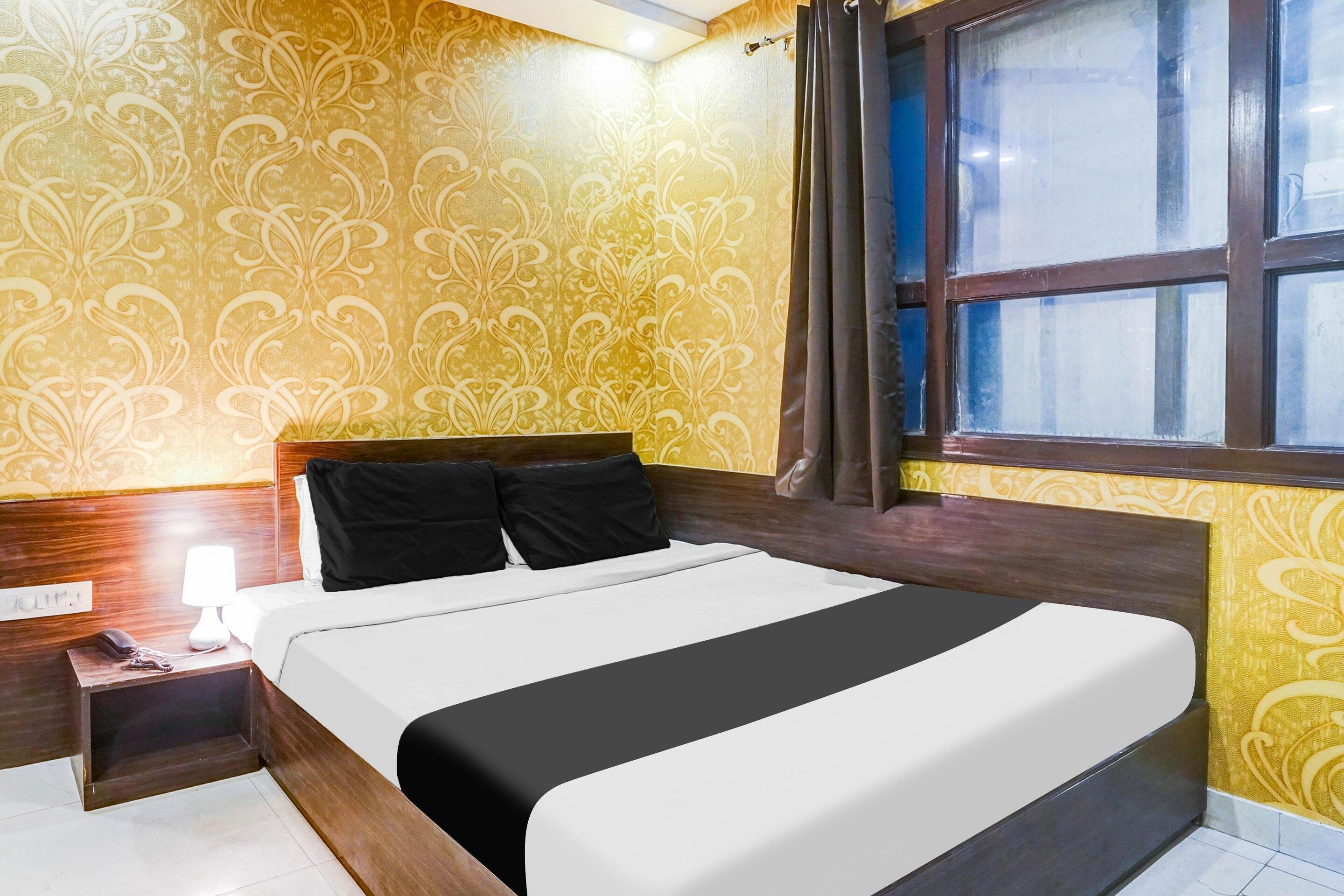 Quarto de hotel com cama, mesa de cabeceira, luminária e janela com cortinas.