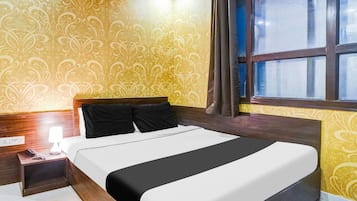 Quarto de hotel com cama, mesa de cabeceira, luminária e janela com cortinas.