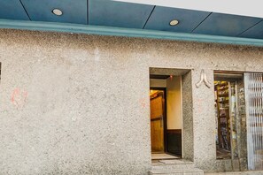 Fachada de edificio con revestimiento de piedra texturizada, un acceso a un espacio interior y una puerta metálica con símbolo de mano.