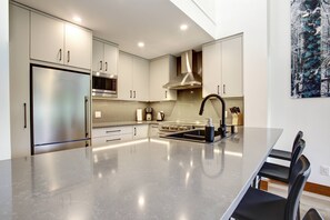A modern kitchen with stainless steel appliances, a large island with a black faucet, and a painting of a forest on the wall. - Alpine Greens 5 Star renovated townhouse (Whistler)